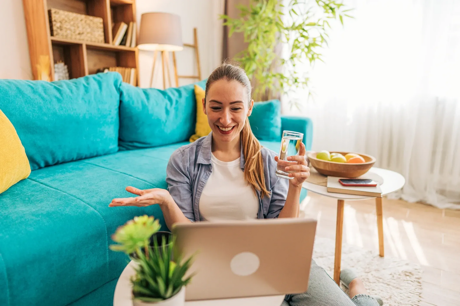 Woman using a laptop in a living room with a blue couch and plants.